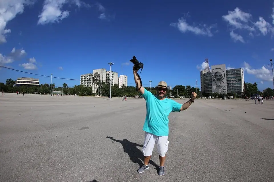 Marcelo levanta su cámara con alegría en una plaza amplia bajo el cielo azul